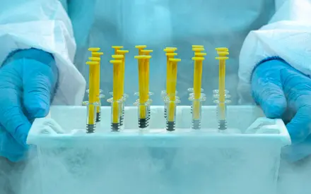 The true cost of cold storage failures - Scientist wearing protective gloves and lab suit holding frozen medical syringes in a cold storage container with visible frost and vapor.